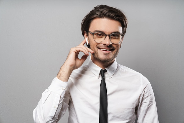 Portrait of a handsome smiling young businessman wearing suit standing isolated over gray wall, listening to music with wireless earphones