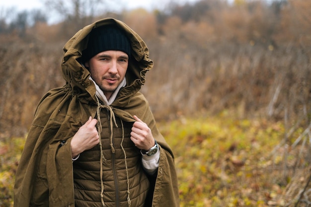 Portrait of handsome traveler man wearing green raincoat tent standing in thicket of bushes in cold overcast day and looking at camera