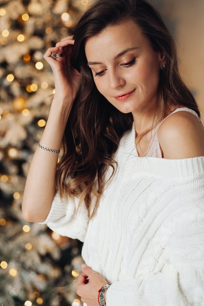 Portrait of handsome woman in fashion white dress smiles near the christmas tree at home