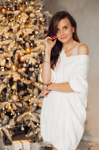 Portrait of handsome woman in white dress smiles near the christmas tree at home