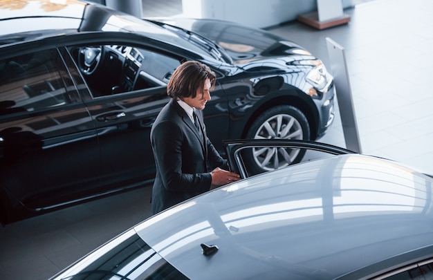 Portrait of handsome young businessman in black suit and tie indoors near modern car.