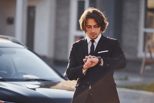 Portrait of handsome young businessman in black suit and tie outdoors near modern car in the city.