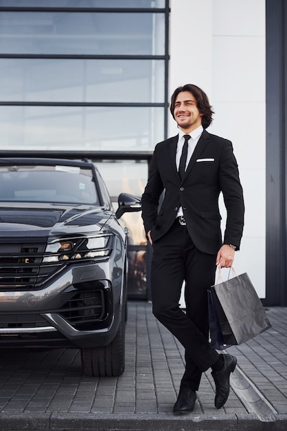 Portrait of handsome young businessman in black suit and tie outdoors near modern car and with shopping bags.