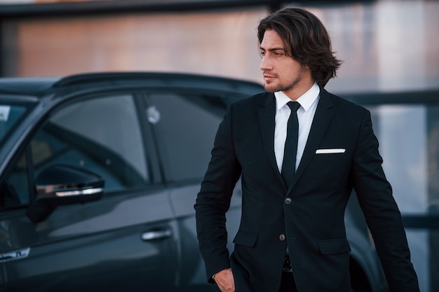 Portrait of handsome young businessman in black suit and tie outdoors near modern car.