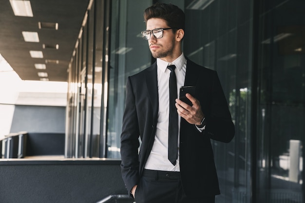 Portrait of handsome young businessman dressed in formal suit standing outside glass building, and holding mobile phone