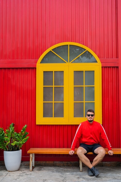 Portrait of a handsome young man dressed in a red jacket and black shorts posing against the background of a red building with a yellow window and potted flowers