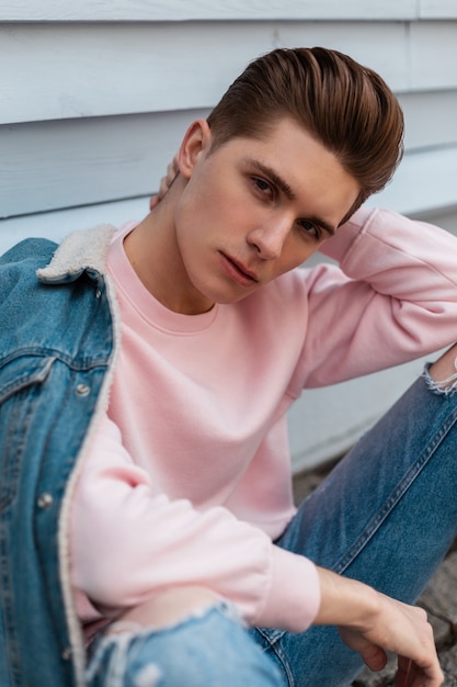 Portrait handsome young man model in trendy pink sweatshirt in fashion denim blue youth jacket near white wall outdoors. Urban guy in stylish jeans casual wear from new collection sit on street.