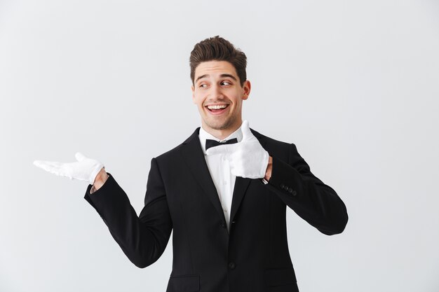 Portrait of a handsome young man waiter wearing tuxedo and gloves standing isolated over white wall, presenting copy space