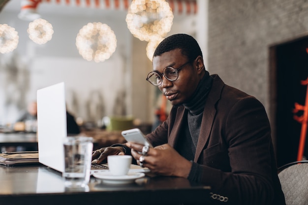 Portrait of happy african businessman using phone while working on laptop in a restaurant.