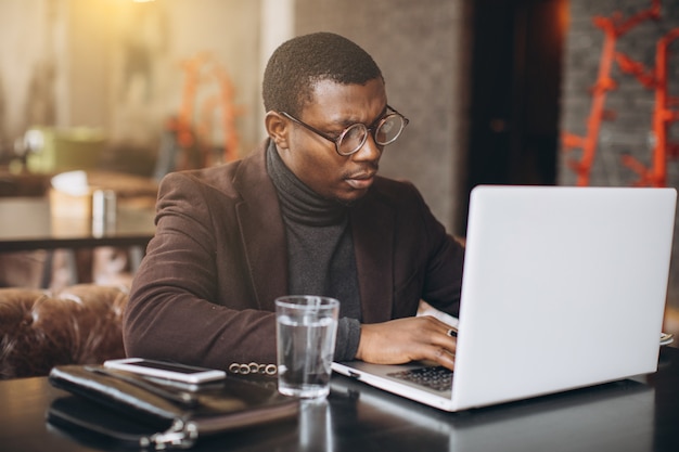 Portrait of happy african businessman using phone while working on laptop in a restaurant.