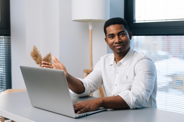 Portrait of happy African male entrepreneur using laptop computer and friendly looking at camera while enjoying work sitting at desk in modern office room