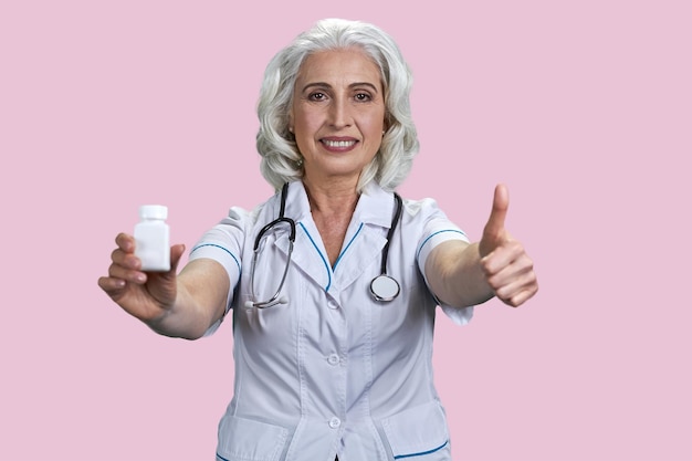 Portrait of happy aged female doctor shows medicine bottle and thumb up advertising pills bottle iso