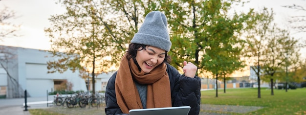 Portrait of happy asian girl sits on bench looks at digital tablet screen and cheers triumphs wins