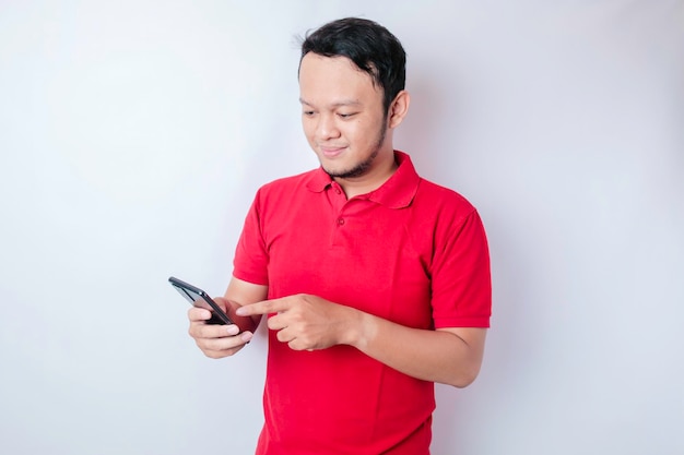 A portrait of a happy Asian man is smiling and holding his smartphone wearing a red tshirt isolated by a white background