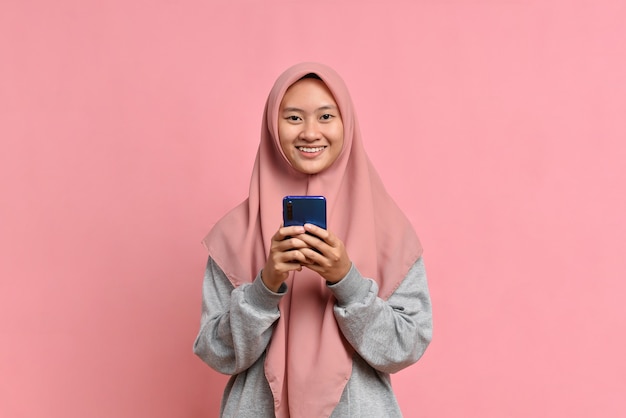 Portrait happy asian muslim woman feeling happiness and looking camera holding smartphone on pink background. Cute asian muslim girl smiling wearing casual hijab and connect internet shopping online