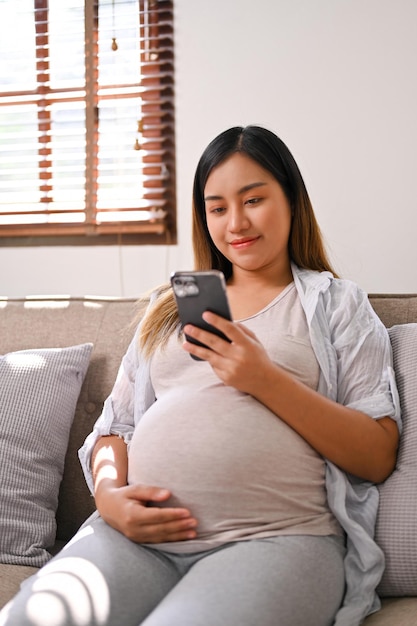 Portrait of a happy Asian pregnant woman using her phone while relaxing on sofa in living room