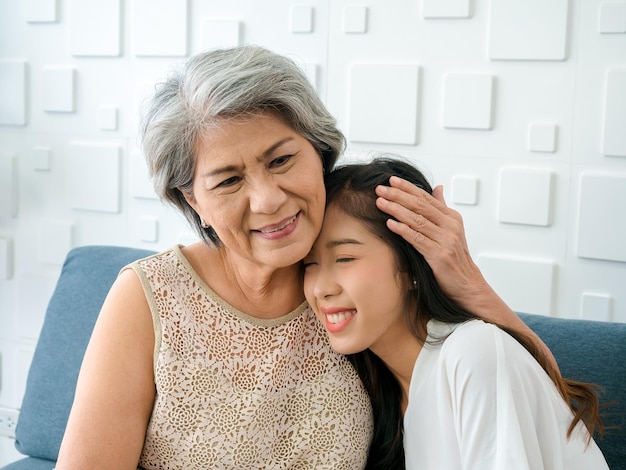 Portrait of happy Asian senior mother white hair embracing her beautiful daughter with closing eyes with love care comfort and smile while sit on sofa on white background in living room at home