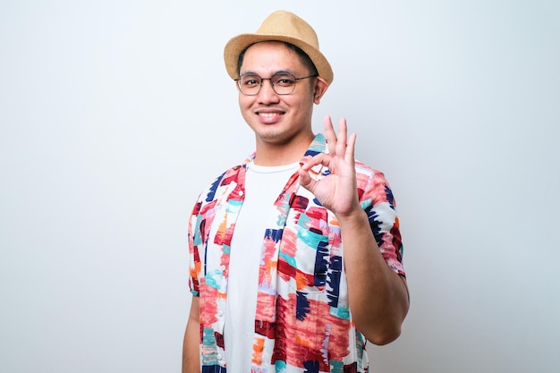 Portrait of happy Asian tourist in casual shirt bucket hat and trendy sunglasses standing and doing ok gesture with finger Traveling holiday trip summer vacation concept