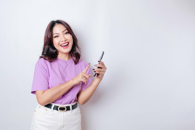 A portrait of a happy Asian woman is smiling and holding her smartphone wearing a lilac purple tshirt isolated by a white background