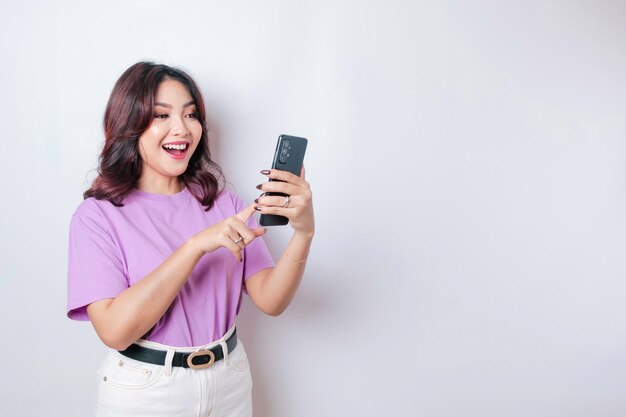 A portrait of a happy Asian woman is smiling and holding her smartphone wearing a lilac purple tshirt isolated by a white background