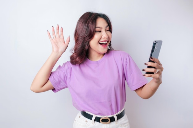 A portrait of a happy Asian woman is smiling and holding her smartphone wearing a lilac purple tshirt isolated by a white background