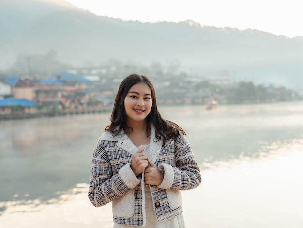 Portrait of happy Asian woman in winter jacket with fog rising on the lake view in the morning at Ban Rak Thai village, Mae Hong Son in Thailand.