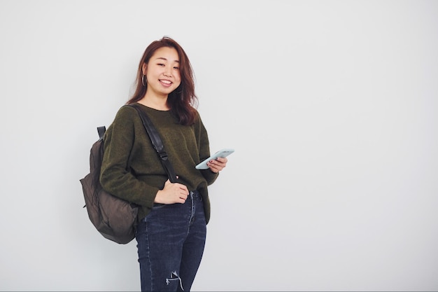 Portrait of happy asian young girl with backpack and phone that standing indoors in the studio against white background.