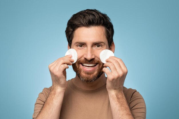 Portrait of happy bearded man holding cotton pads under eyes and smiling at camera standing over blue studio background