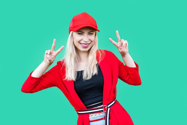 Portrait of happy beautiful blond young hipster style woman in red blouse and cap, standing with victory sign and looking at camera with toothy smile. indoor studio shot, isolated on green background