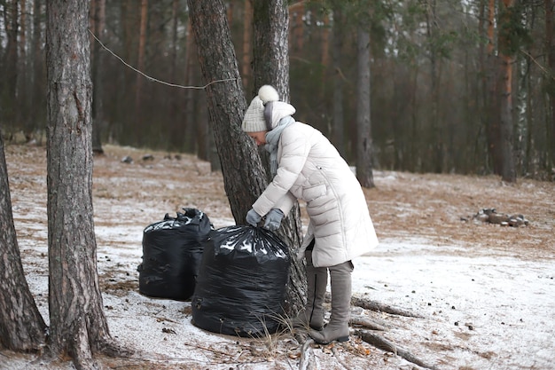 Portrait of happy beautiful elderly senior retired woman in age in forest or park at winter cold day smiling picking up garbage bag Side viewThe concept of environment protection and volunteering