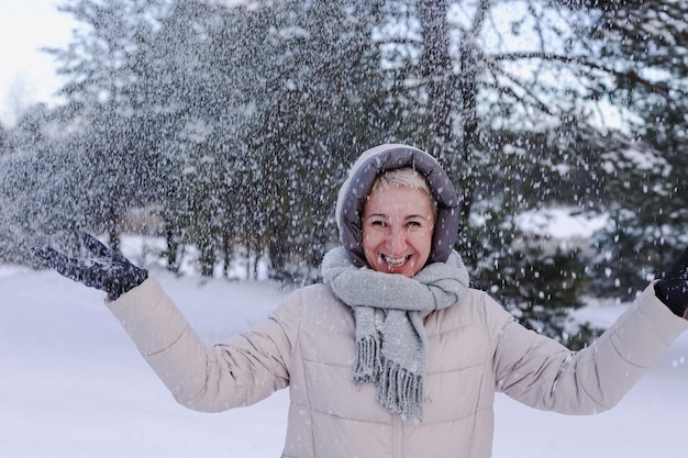 Portrait of happy beautiful elderly senior retired woman in age is playing having fun with snow outdoors in forest or park at winter cold day smiling enjoy weather