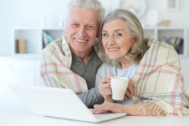 Portrait of happy beautiful senior couple with blanket using laptop at home