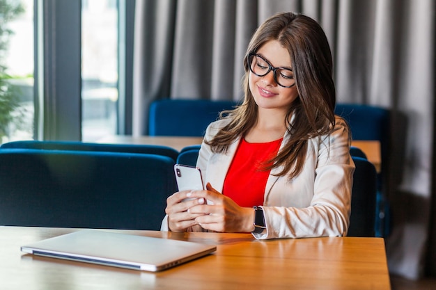 Portrait of happy beautiful stylish brunette young woman in glasses sitting holding her mobile smart phone and looking with toothy smile indoor studio shot cafe office background