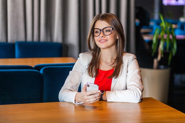 Portrait of happy beautiful stylish brunette young woman in glasses sitting with mobile smart phone and looking away with toothy smile and dreaming indoor studio shot cafe office background