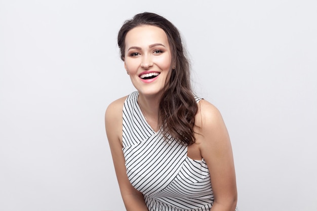 Portrait of happy beautiful young brunette woman with makeup and striped dress standing and looking at camera with toothy smile and excited face. indoor studio shot, isolated on grey background.