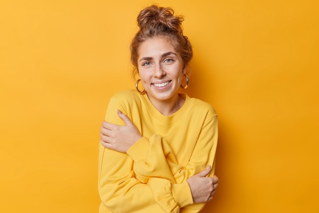 Portrait of happy beautiful young woman embraces herself smiles tenderly feels comfortable in casual jumper has combed hair poses against yellow background People lifestyle happiness concept