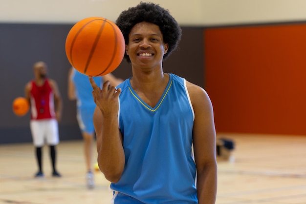 Portrait of happy biracial male basketball player doing trick with basketball over teammates at gym. Sport, activity and lifestyle, unaltered.