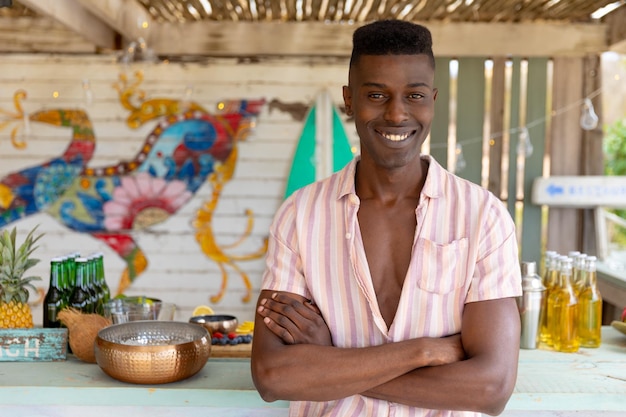 Portrait of happy biracial man smiling standing by counter at sunny beach bar, unaltered. Small business, summer, relaxation, refreshment and vacation.