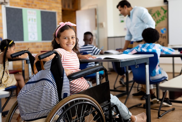 Portrait of happy biracial school girl sitting in wheelchair in class at elementary school