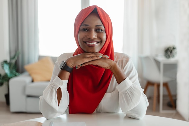 Portrait of happy black muslim lady in casual outfit and hijab posing at workplace at home smiling at camera