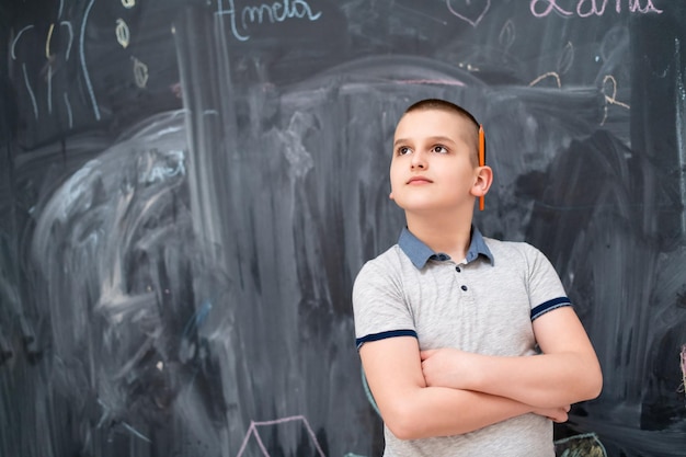 portrait of happy boy with orange wooden pen behind the ear standing in front of black chalkboard