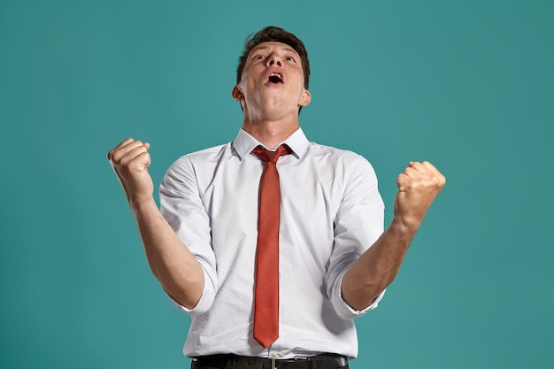 Portrait of a happy brunet guy with brown eyes, wearing in a classic white shirt and red tie. He is acting like has won something posing in a studio against a blue background. Concept of gesticulation