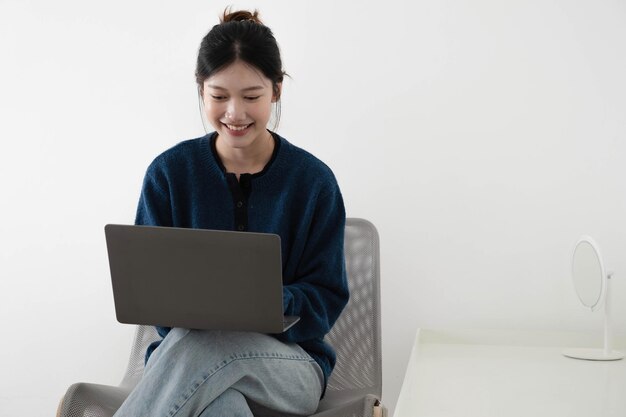 portrait of a happy casual asian woman holding laptop computer while sitting on a chair over white background