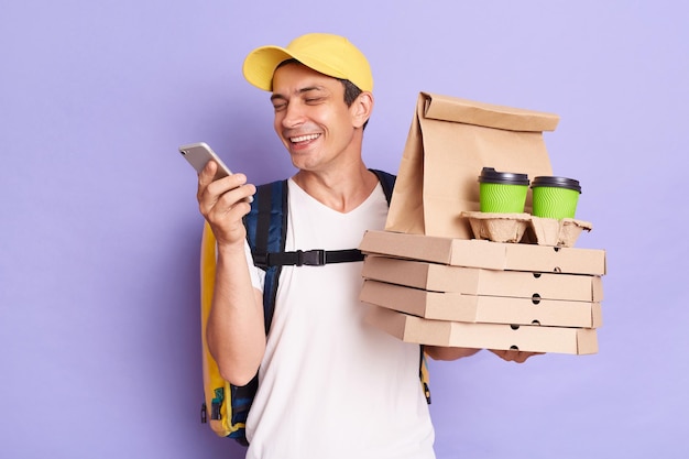 Portrait of happy Caucasian delivery man with food and drinks standing isolated over purple background holding mobile phone checking address of delivering