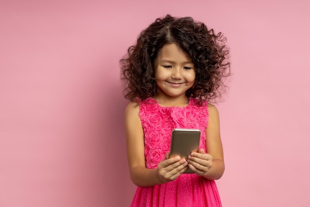 Portrait of happy caucasian little girl with curly hairstyle, wearing dress, holding mobile phone, watching videos, looking at screen with joyful smile. People, technology, children, internet concept.