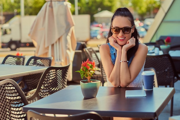 Portrait of a happy charming brunette girl wearing trendy clothes is enjoying summer day while sitting on a terrace in outdoors cafe, looking at the camera.