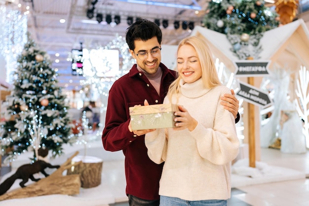 Portrait of happy cheerfully blonde woman receiving gift box from loving husband standing in hall of celebrate shopping mall in Christmas eve