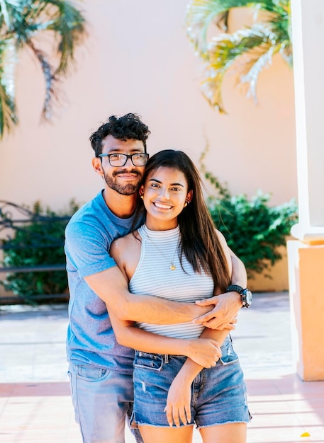 Portrait of a happy couple man hugging his girlfriend from behind Portrait of a young couple man hugging his girlfriend from behind outside Happy couple embracing looking at the camera