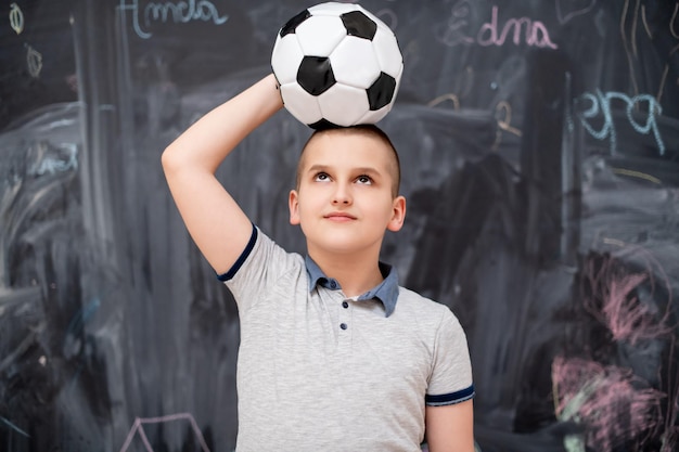 portrait of happy cute boy having fun holding a soccer ball on his head while standing in front of black chalkboard