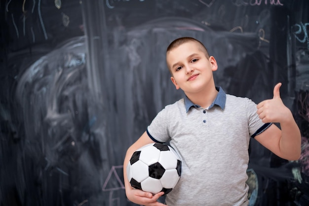 portrait of happy cute boy having fun holding a soccer ball while standing in front of black chalkboard